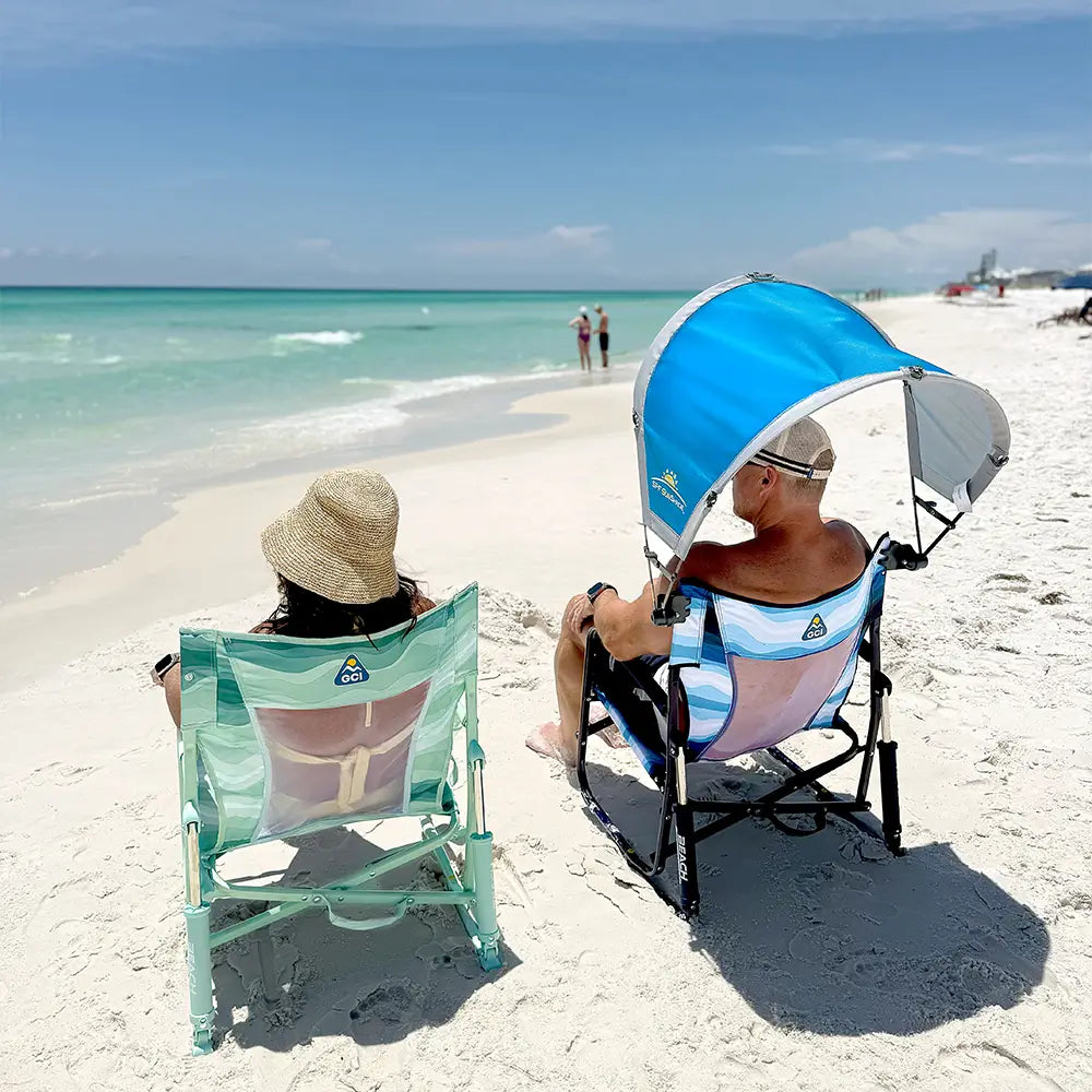 A man sitting in a beach rocker on the beach with the saybrook blue sunshade accessory attached to the chair.